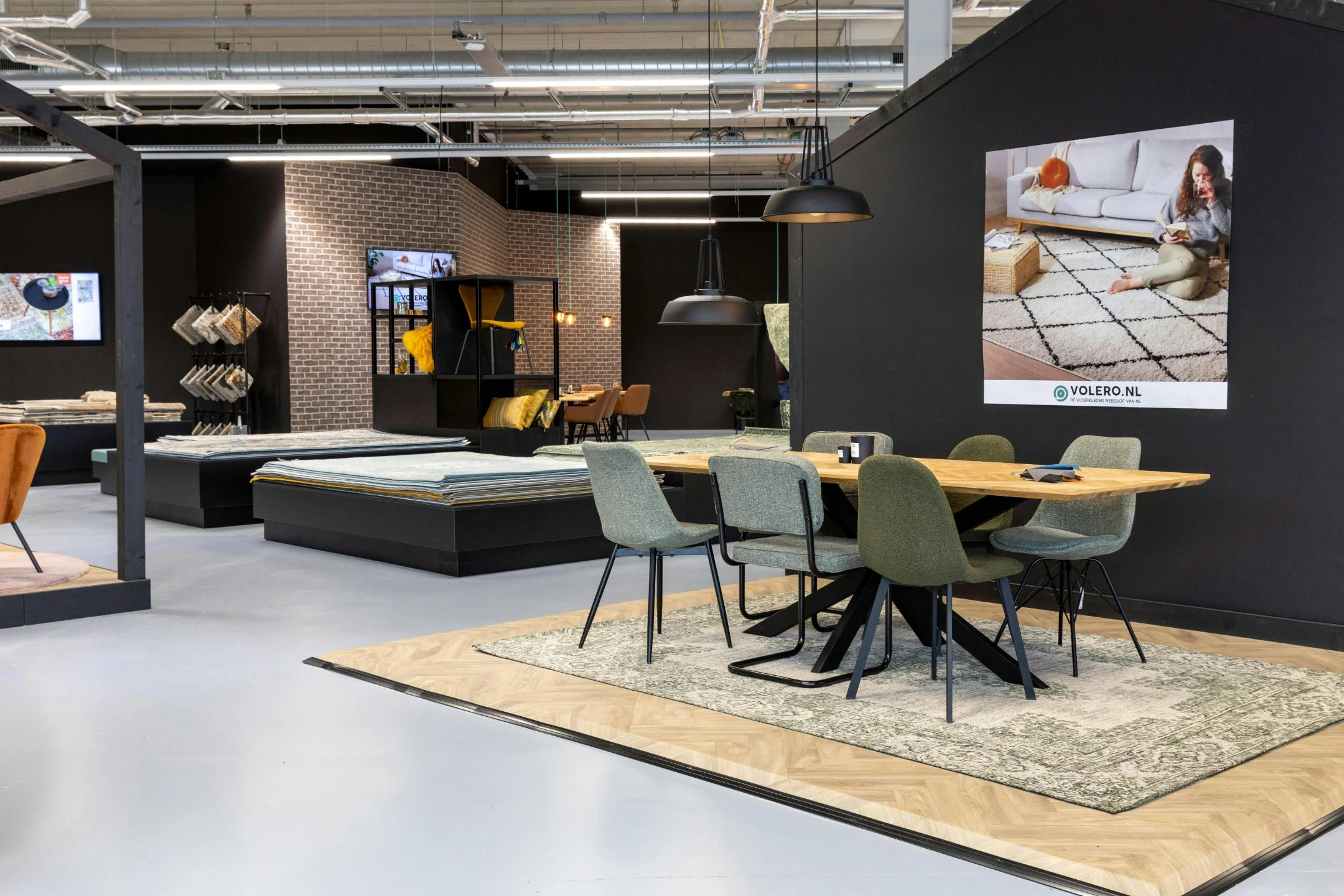 Dining table setting on a rug beside a VOLERO.NL wall poster and rug sample displays in the Amsterdam showroom