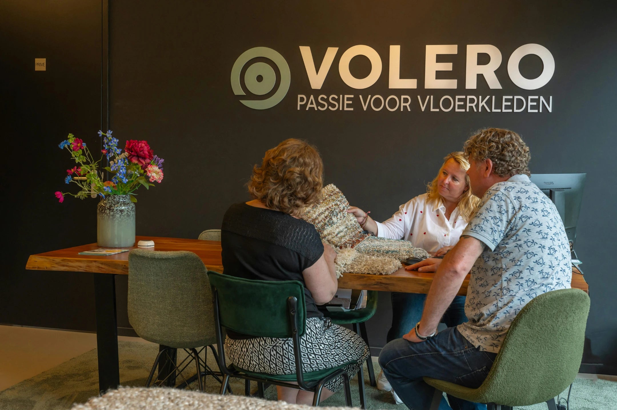 Customers reviewing rug samples with a Volero adviser at a wooden table beneath the Volero Passie voor Vloerkleden wall logo in the Lekkerkerk showroom