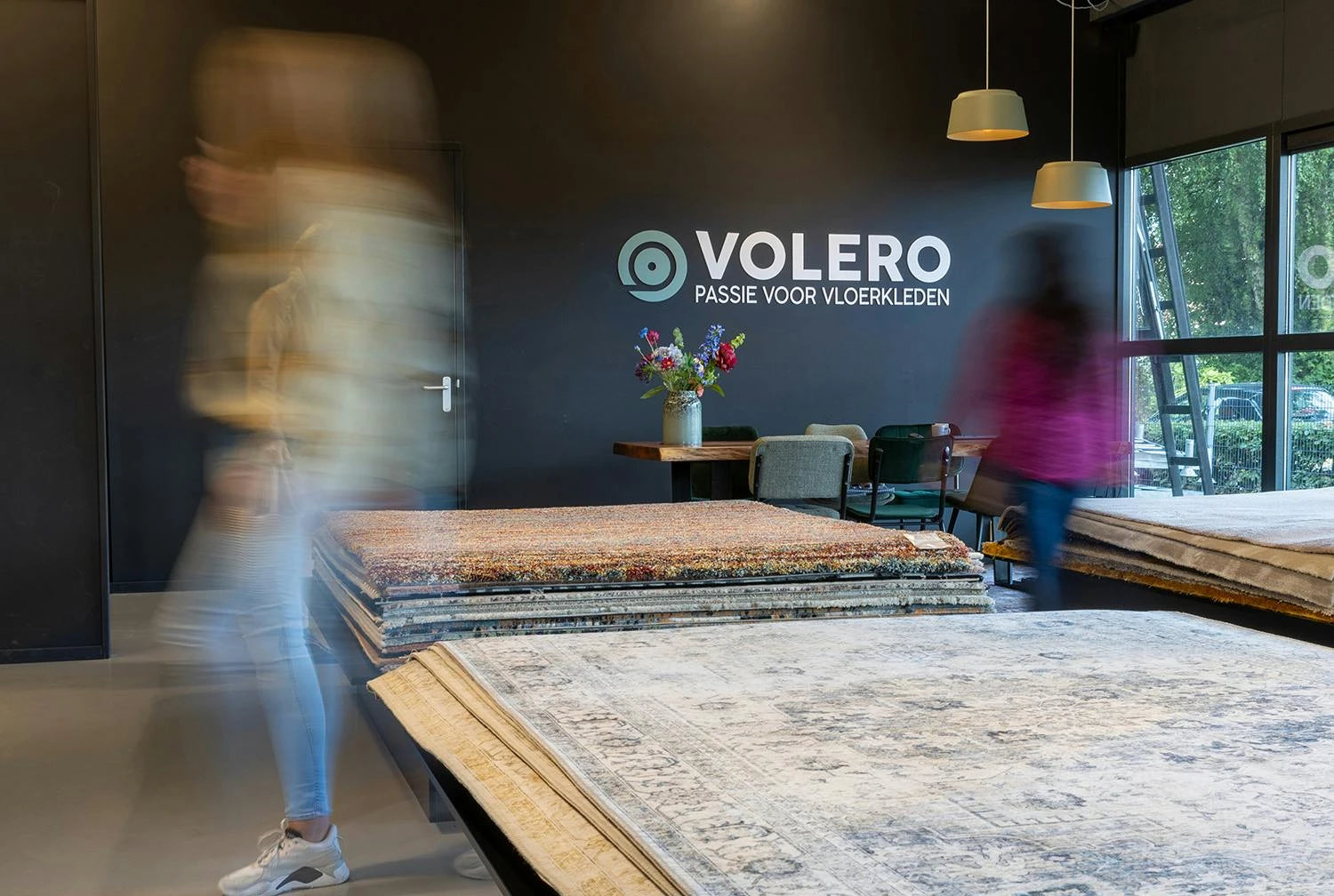 Visitors walking past stacked rug displays in the Volero Lekkerkerk showroom, with the Volero Passie voor Vloerkleden logo on the back wall